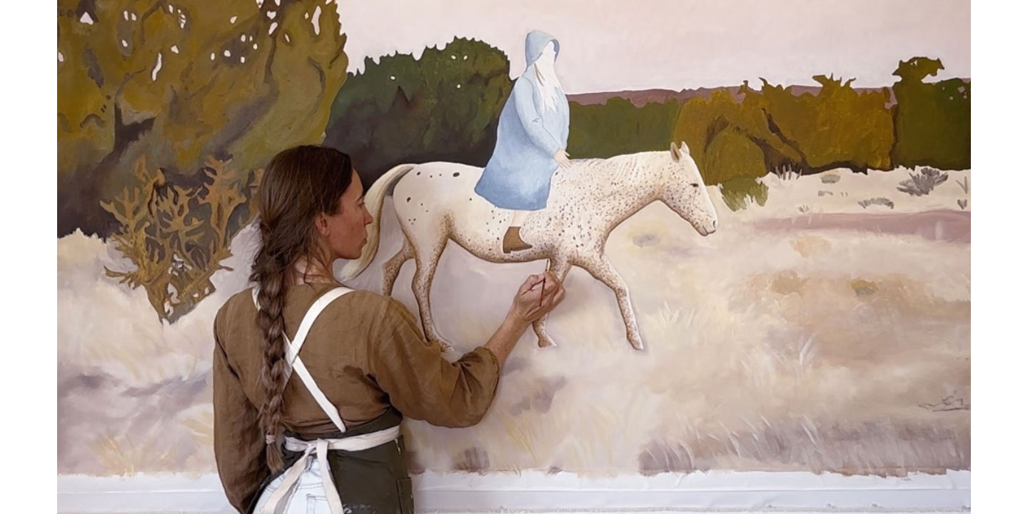 A woman, Stella Maria Baer, is on her knees against a wall, painting a white horse with a girl wearing a blue cloak riding on it and a multicolored sunset in the background.